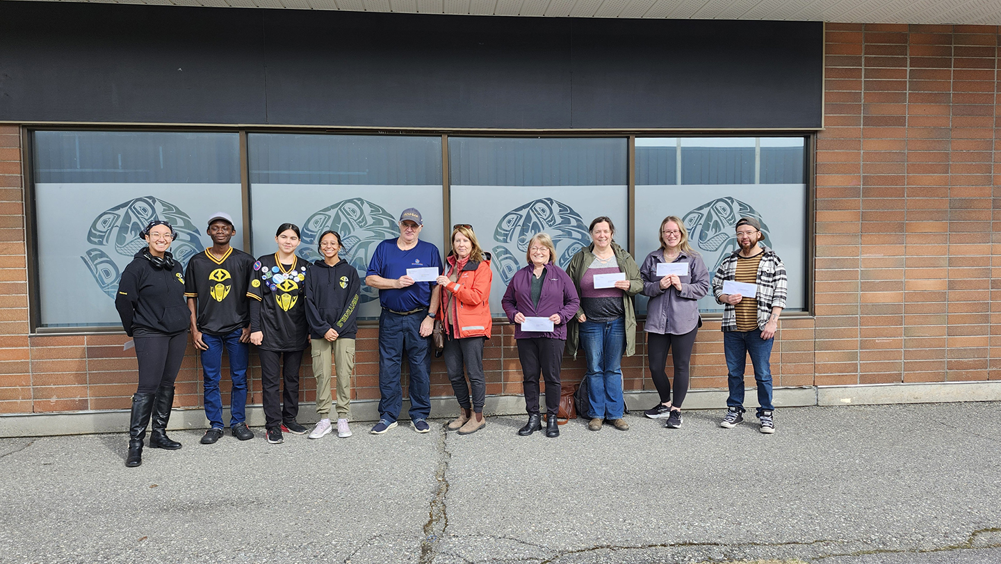 Ten recipients stand in front of a glass windown showing a cheque from the Kitimat Community foundation